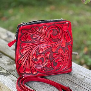 Peut inclure: Un sac &agrave; bandouli&egrave;re en cuir rouge avec un motif floral complexe. Le sac a une fermeture &agrave; glissi&egrave;re et une bandouli&egrave;re r&eacute;glable.