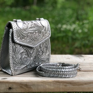 Pode incluir: Uma bolsa de m&atilde;o de couro prateada com um design floral intrincado em relevo na frente. A bolsa tem uma al&ccedil;a de ombro e um fecho de aba.