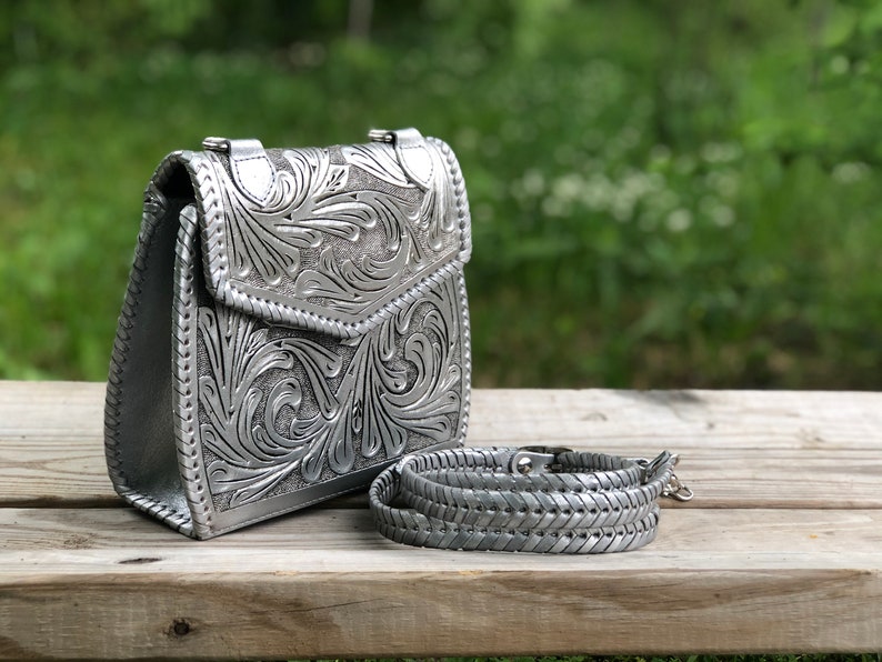 Pode incluir: Uma bolsa de m&atilde;o de couro prateada com um design floral intrincado em relevo na frente. A bolsa tem uma al&ccedil;a de ombro e um fecho de aba.