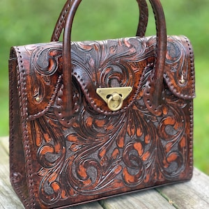 Puede incluir: Un bolso de mano de cuero marr&oacute;n con intrincados detalles florales y un cierre de lat&oacute;n. El bolso tiene dos asas superiores.