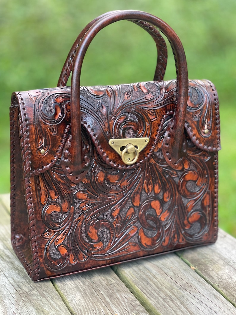 Puede incluir: Un bolso de mano de cuero marr&oacute;n con intrincados detalles florales y un cierre de lat&oacute;n. El bolso tiene dos asas superiores.