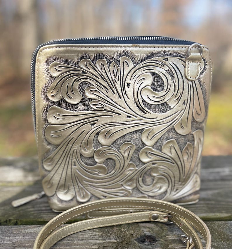 Peut inclure: Un sac &agrave; bandouli&egrave;re en cuir dor&eacute; avec un motif floral complexe. Le sac a une fermeture &agrave; glissi&egrave;re et une longue bandouli&egrave;re r&eacute;glable.
