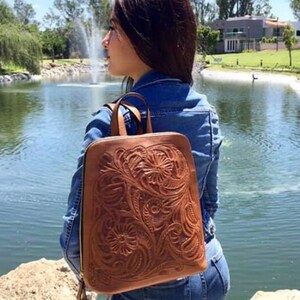 May include: A brown leather backpack with intricate floral tooling. The backpack has a zipper closure and adjustable straps. The backpack is being worn by a person standing near a pond.