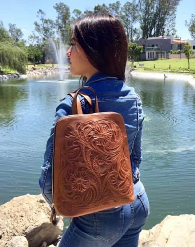 May include: A brown leather backpack with intricate floral tooling. The backpack has a zipper closure and adjustable straps. The backpack is being worn by a person standing near a pond.