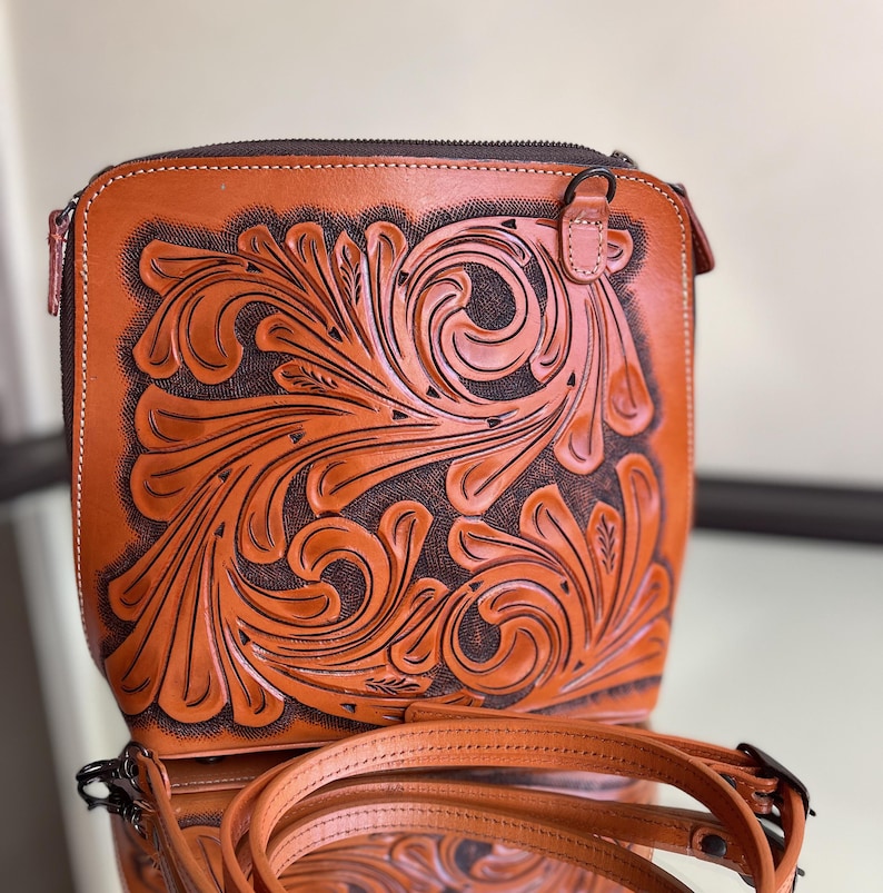 Peut inclure: Un sac &agrave; bandouli&egrave;re en cuir marron avec un travail floral complexe. Le sac a une fermeture &agrave; glissi&egrave;re et une bandouli&egrave;re r&eacute;glable.