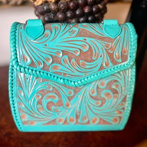 Pode incluir: Uma bolsa de couro azul turquesa com um design floral em relevo. A bolsa tem um fecho de aba e uma guarni&ccedil;&atilde;o tran&ccedil;ada.