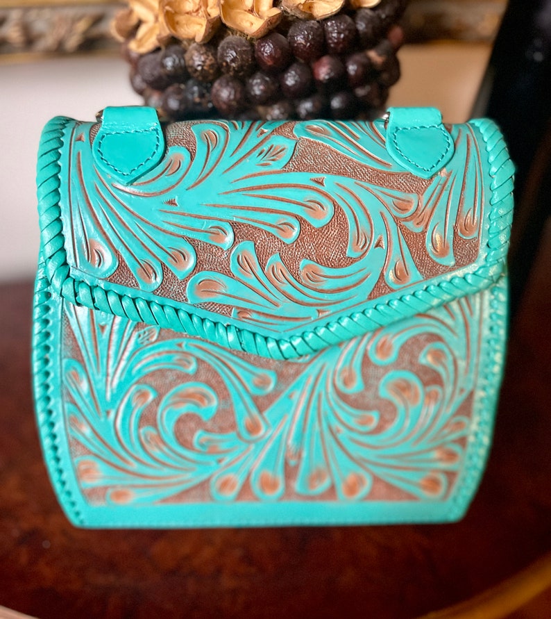 Pode incluir: Uma bolsa de couro azul turquesa com um design floral em relevo. A bolsa tem um fecho de aba e uma guarni&ccedil;&atilde;o tran&ccedil;ada.