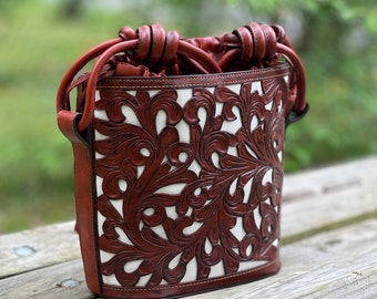 Peut inclure: Un sac seau en cuir marron avec une doublure en toile blanche et des découpes florales complexes. Le sac a une fermeture à cordon et une poignée supérieure.