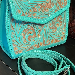 Pode incluir: Uma bolsa de couro turquesa com detalhes florais intrincados e uma al&ccedil;a tran&ccedil;ada combinando. A bolsa tem fecho de aba e uma fivela prateada.