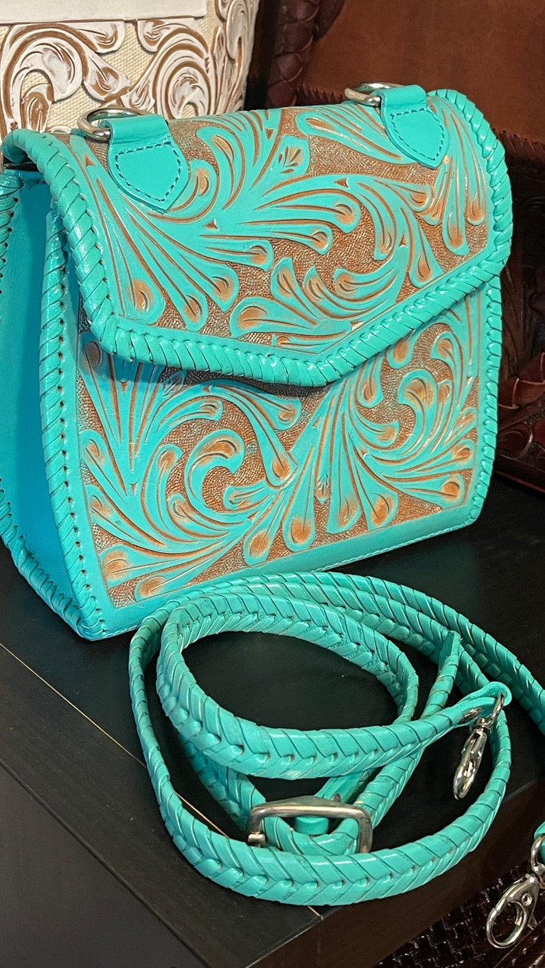 Pode incluir: Uma bolsa de couro turquesa com detalhes florais intrincados e uma al&ccedil;a tran&ccedil;ada combinando. A bolsa tem fecho de aba e uma fivela prateada.