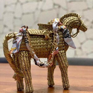 Pu&ograve; includere: Una borsa a forma di cavallo intrecciata color oro, con una sella e una tracolla a catena. Il cavallo ha una sciarpa decorativa legata intorno al collo e alla sella. La borsa &egrave; realizzata in un materiale simile alla paglia.