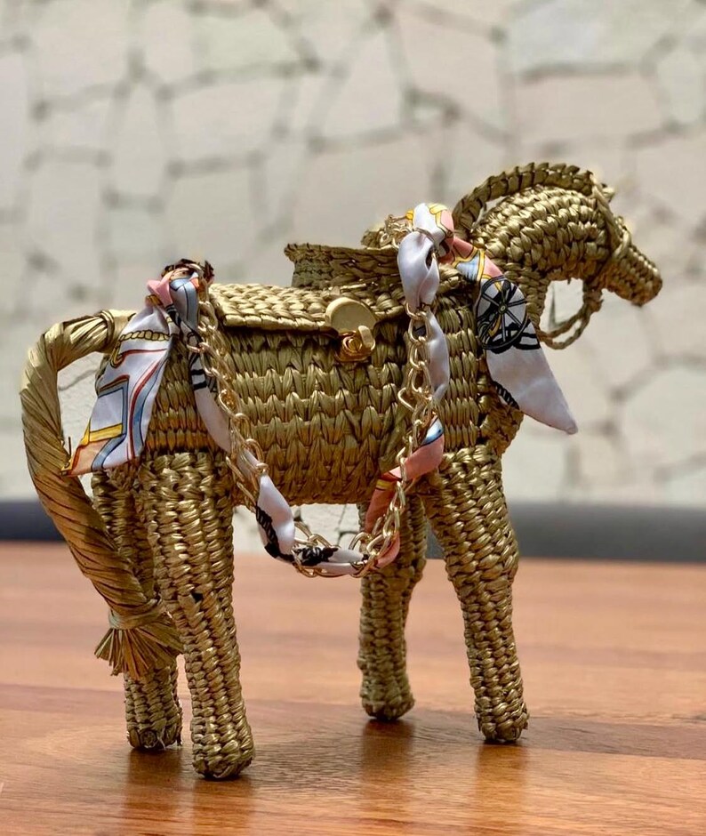 Pu&ograve; includere: Una borsa a forma di cavallo intrecciata color oro, con una sella e una tracolla a catena. Il cavallo ha una sciarpa decorativa legata intorno al collo e alla sella. La borsa &egrave; realizzata in un materiale simile alla paglia.