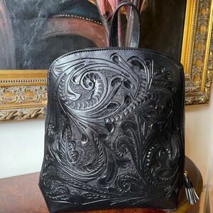 May include: Black leather backpack with an intricate floral pattern embossed on the front. The bag has a top handle and a zipper closure.