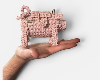 Può includere: Una borsa a forma di maiale rosa intrecciata con una piccola chiusura color oro. La borsa ha un manico intrecciato e una piccola etichetta rettangolare che dice "LA fourmi". Il maiale è tenuto nella mano di una persona.