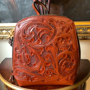 May include: A brown leather backpack with intricate floral tooling. The backpack has a zipper closure and a tassel pull.