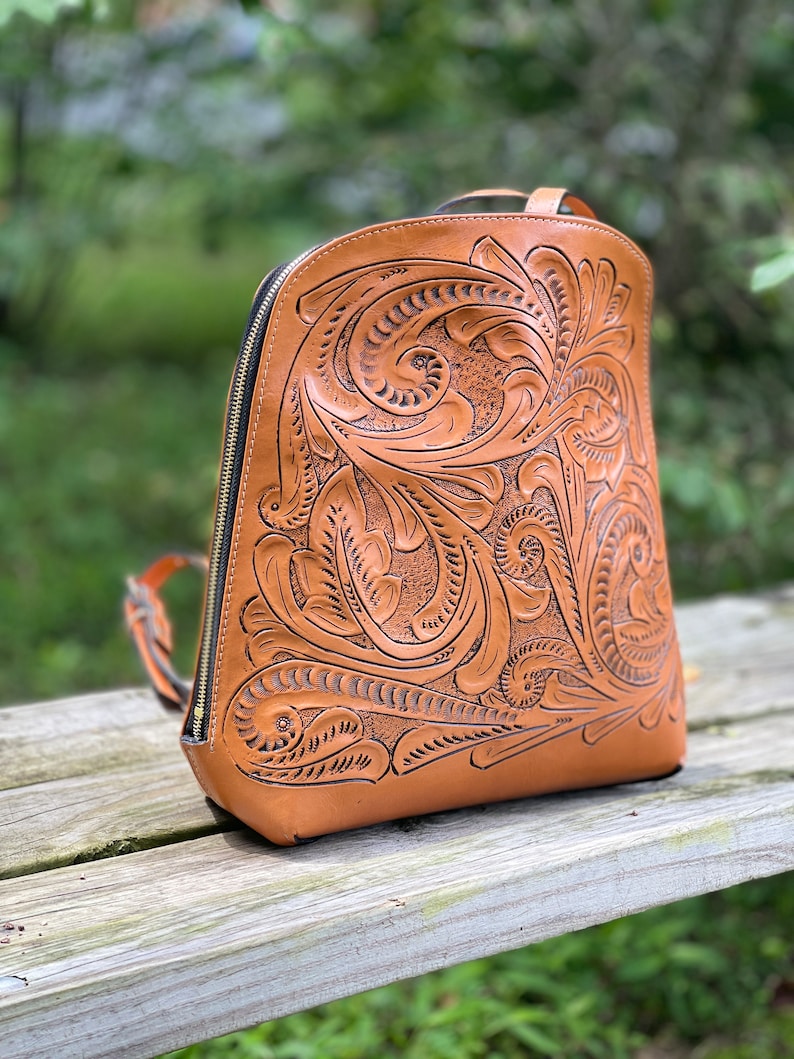 May include: A brown leather backpack with intricate floral tooling. The backpack has a zipper closure and adjustable straps.
