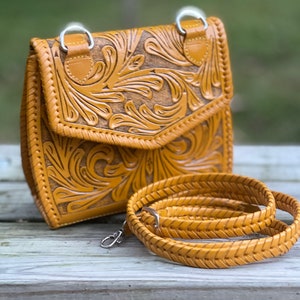 Pode incluir: Uma bolsa de ombro de couro amarelo mostarda com detalhes florais intrincados. A bolsa tem fecho de aba e uma al&ccedil;a tran&ccedil;ada.