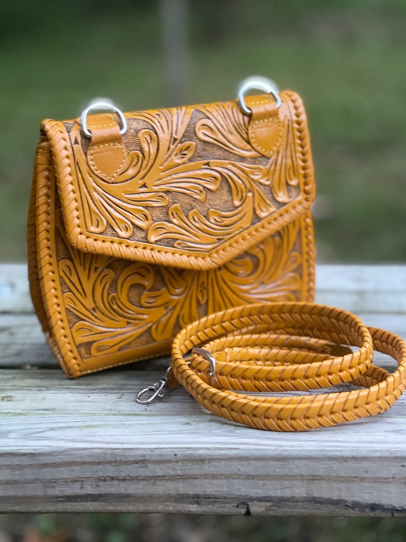 Pode incluir: Uma bolsa de ombro de couro amarelo mostarda com detalhes florais intrincados. A bolsa tem fecho de aba e uma al&ccedil;a tran&ccedil;ada.