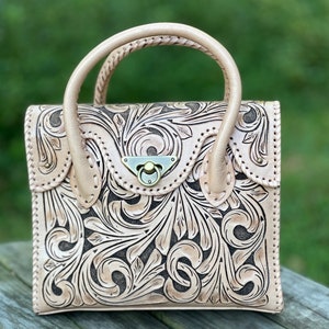Puede incluir: Un bolso de mano de cuero marr&oacute;n claro con un dise&ntilde;o floral tallado. El bolso tiene un asa superior y un cierre de broche.