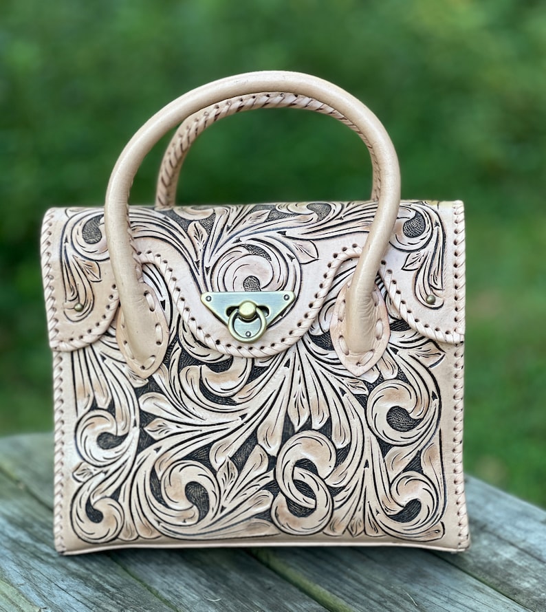 Puede incluir: Un bolso de mano de cuero marr&oacute;n claro con un dise&ntilde;o floral tallado. El bolso tiene un asa superior y un cierre de broche.