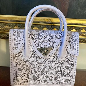 Puede incluir: Un bolso de mano de cuero lavanda con intrincados dise&ntilde;os florales tallados. El bolso tiene un asa superior y un cierre de broche.
