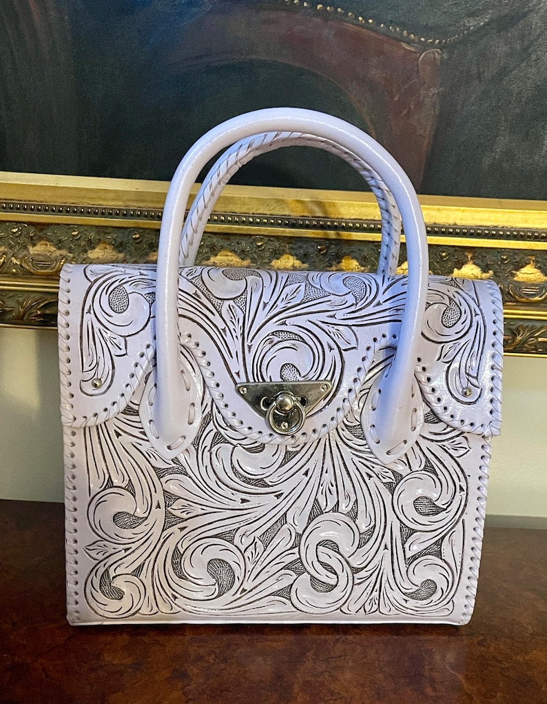 Puede incluir: Un bolso de mano de cuero lavanda con intrincados dise&ntilde;os florales tallados. El bolso tiene un asa superior y un cierre de broche.
