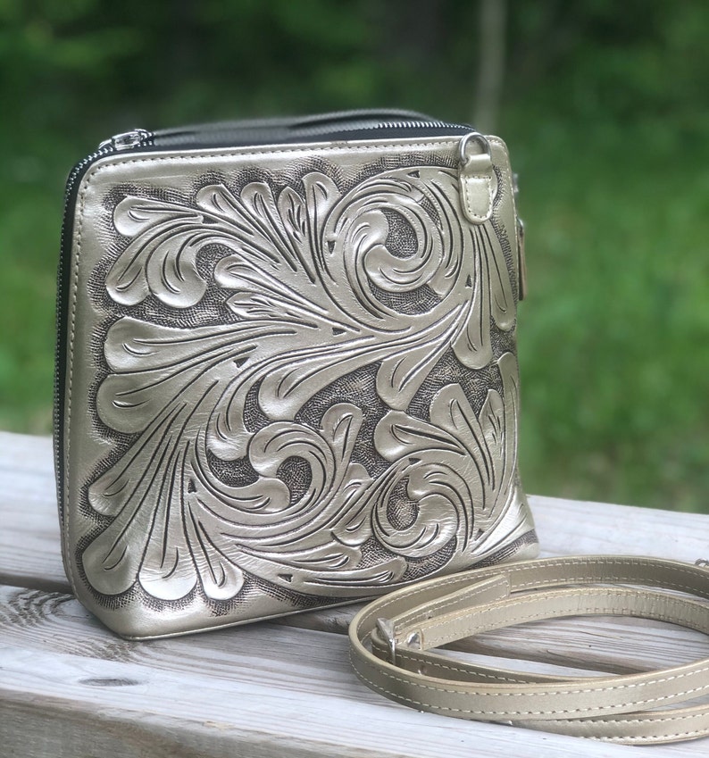 Peut inclure: Un sac &agrave; bandouli&egrave;re en cuir argent&eacute; avec un motif floral complexe. Le sac a une fermeture &agrave; glissi&egrave;re et une longue bandouli&egrave;re r&eacute;glable.