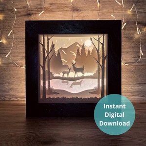 May include: A black framed shadow box featuring a paper cut design of a forest scene with two deer, a lake, and mountains. The scene is illuminated from behind, creating a glowing effect.