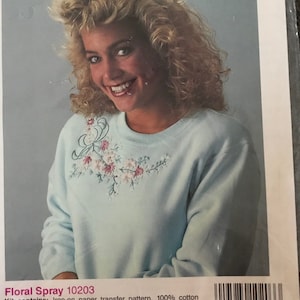 May include: A blue sweatshirt with a floral embroidery design. The embroidery is in shades of pink, white, and green. The sweatshirt is part of an Iron-On Fashions kit, which includes a transfer pattern, 100% cotton floss, rhinestones, a needle, and easy-to-follow instructions. The kit is made by Golden Bee Stitchery, Inc. and has the product number 10203.