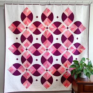 May include: A large, geometric quilt with a white background and a repeating pattern of pink and purple shapes. The design features circles, diamonds, and petal-like forms. The quilt is hanging on a white rod against a white wall.