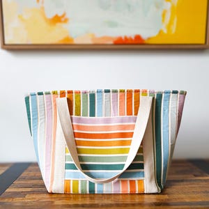 Pode incluir: Uma sacola colorida com listras verticais em tons de azul, verde, laranja e rosa. A sacola tem um bolso creme e alças combinando. A sacola está em uma mesa de madeira.