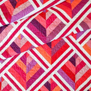 May include: A quilt with a geometric pattern in shades of pink, red, and purple. The quilt is made of squares and diamonds, and the fabric is quilted with a wavy pattern.