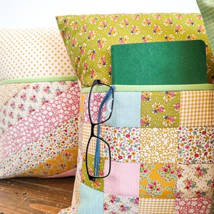 May include: A patchwork pillow with a green book and glasses tucked into a pocket. The pillow is made of various floral and geometric fabrics in shades of pink, yellow, green, and blue.