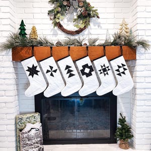 May include: A fireplace mantel decorated for Christmas. Six white stockings with black geometric patterns hang above the fireplace. A wreath with pinecones and a black and white striped bow is above the stockings. Small Christmas trees and greenery complete the festive scene.