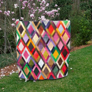 Op de afbeelding: Een kleurrijke quilt met een ruitpatroon. De quilt heeft een verscheidenheid aan kleuren, waaronder rood, roze, groen, geel en bruin. De quilt wordt buiten op een grasveld met een boom op de achtergrond getoond.