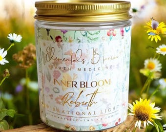 May include: A glass jar with a gold lid, adorned with floral patterns and the words "Elementals Botanica Earth Medicine Inner Bloom Rebirth Vibrational Light." The jar is surrounded by wildflowers and greenery.