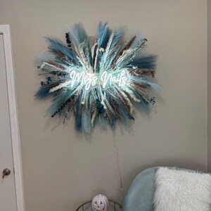 May include: A neon sign reading "Megs Nails" surrounded by blue, white, and brown dried flowers and grasses. The sign is mounted on a light grey wall. A teal armchair and a small table with a pumpkin decoration are in the foreground. The sign is a decorative item.