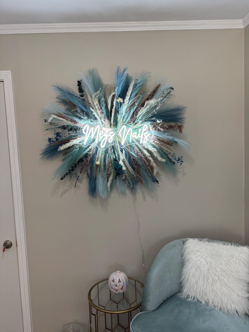 May include: A neon sign reading "Megs Nails" surrounded by blue, white, and brown dried flowers and grasses. The sign is mounted on a light grey wall. A teal armchair and a small table with a pumpkin decoration are in the foreground. The sign is a decorative item.