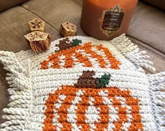 May include: Two crocheted pumpkin coasters with fringe edges. The coasters are white with orange and brown details. A candle with a label that reads "Leaves" is in the background.