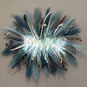 May include: A wall decoration with a neon sign reading "Meg's Nails", encircled by dried flowers and grasses in blue, green, and brown hues. The circular arrangement is mounted on a neutral wall, creating a natural aesthetic.