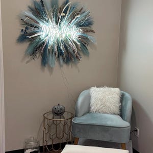 May include: Interior of a nail salon with a light blue velvet chair and a fluffy white cushion. A gold side table and foot spa are also visible. A neon sign reading "Meg's Nails" is mounted on the wall, surrounded by blue and beige decorations.
