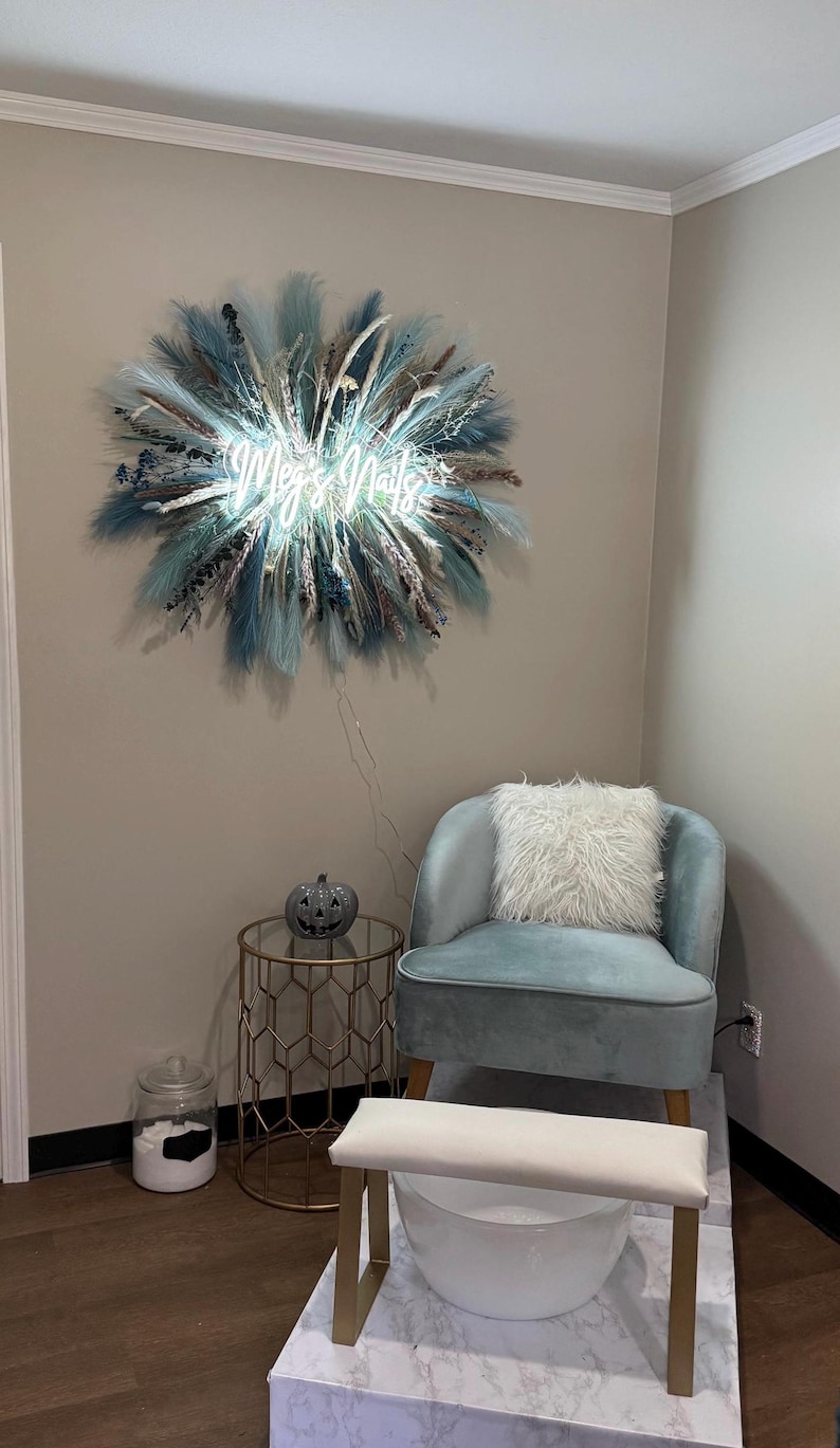 May include: Interior of a nail salon with a light blue velvet chair and a fluffy white cushion. A gold side table and foot spa are also visible. A neon sign reading "Meg's Nails" is mounted on the wall, surrounded by blue and beige decorations.