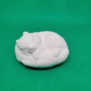 May include: White ceramic figurine of a sleeping cat. The cat is curled up in a ball with its head resting on its paws.