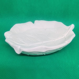 May include: A white, leaf-shaped decorative dish. The dish has detailed veining and a gently curved shape. The background is a solid green color.
