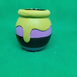 May include: A small, round ceramic pot with a yellow, purple, and black striped design. The pot has a yellow glaze that drips down the sides.