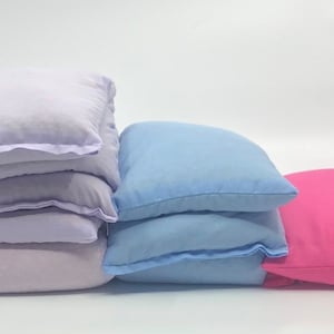 Può includere: Una pila di sei cuscini quadrati lavanda con un bordo viola chiaro, accanto a due cuscini azzurri e un cuscino rosa. I cuscini sono in tessuto morbido e sono disposti su uno sfondo bianco.