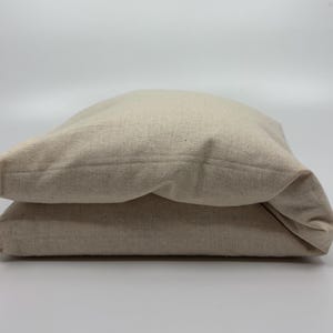 May include: Two beige linen pillow covers are stacked on a white surface. The pillow covers are made of a textured fabric and have a simple, minimalist design. The neutral color and natural material suggest a focus on comfort and simplicity.