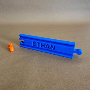 May include: A blue plastic track piece with the name "ETHAN" engraved on it. The track piece has a small orange connector on the side.