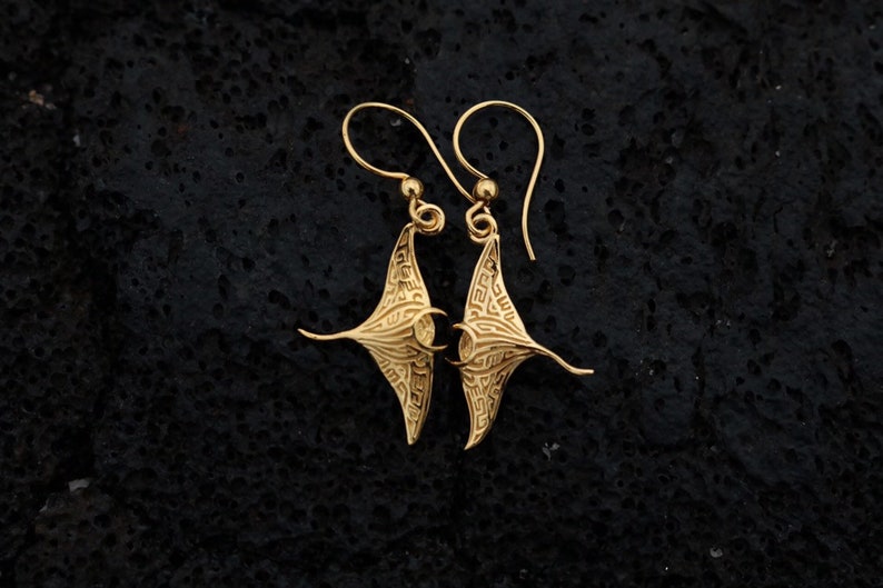 May include: Gold-tone earrings featuring a detailed manta ray design. The earrings have a dangling style with a hook closure.