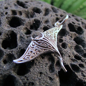 Puede incluir: Un colgante de plata con forma de manta raya con intrincados diseños geométricos. El colgante está sobre una superficie texturizada gris oscuro.
